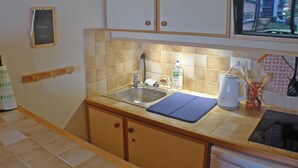 Een keuken met houten kastjes, een spoelbak, een waterkoker en een klein schoolbord aan de muur.