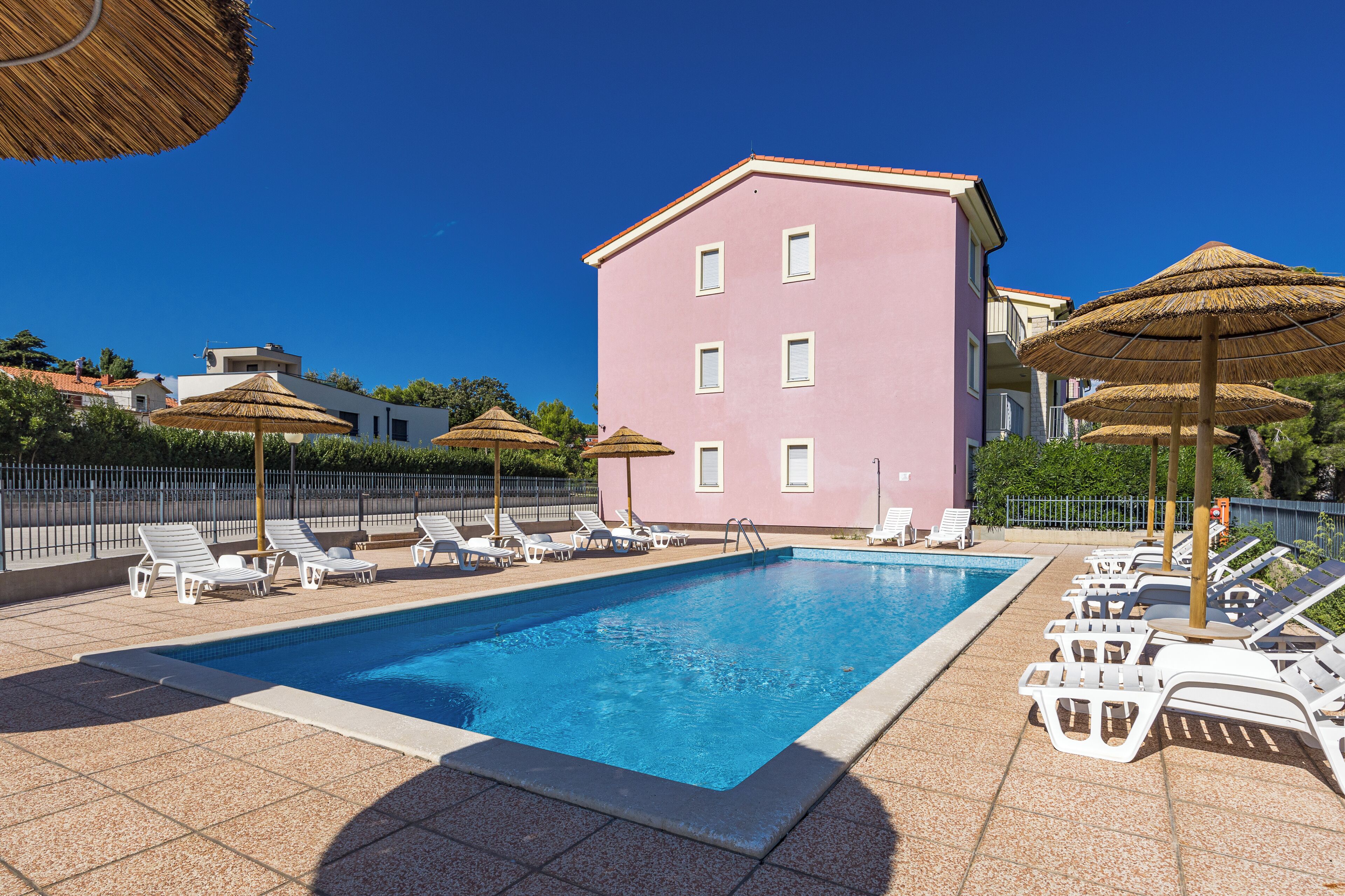 A swimming pool with lounge chairs and umbrellas, surrounded by a pink building.