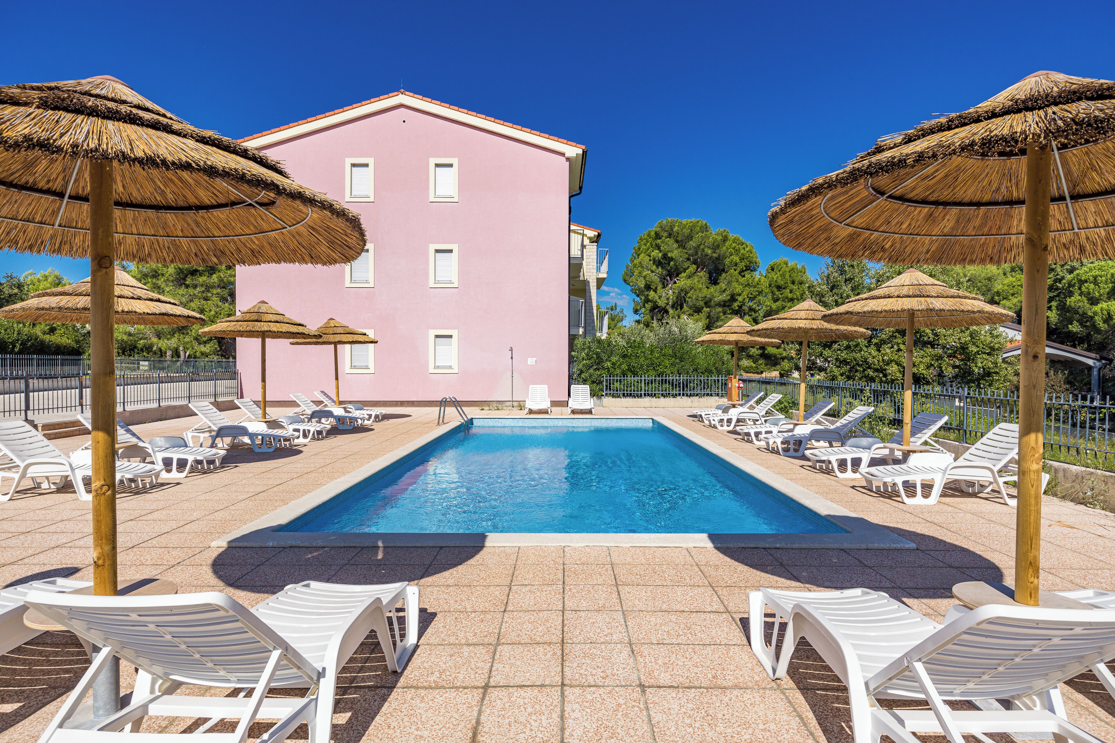Une piscine entourée d’un bâtiment rose, avec des chaises longues blanches et des parasols en chaume.