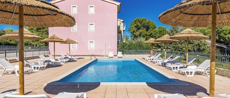 Une piscine entourée d’un bâtiment rose, avec des chaises longues blanches et des parasols en chaume.