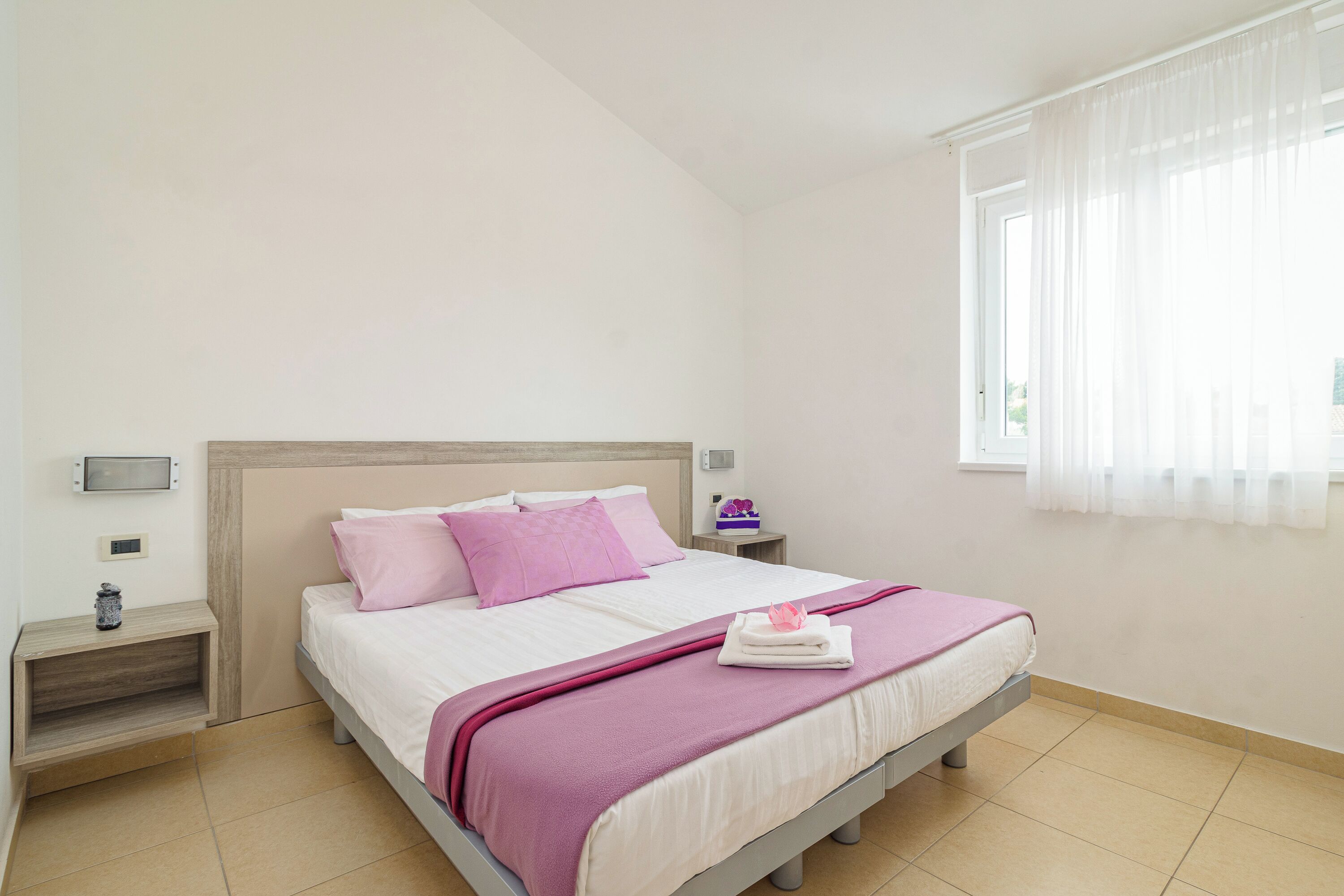 A neatly made bed with a pink blanket and pillows, a bedside table with a lamp, and a window with sheer curtains.