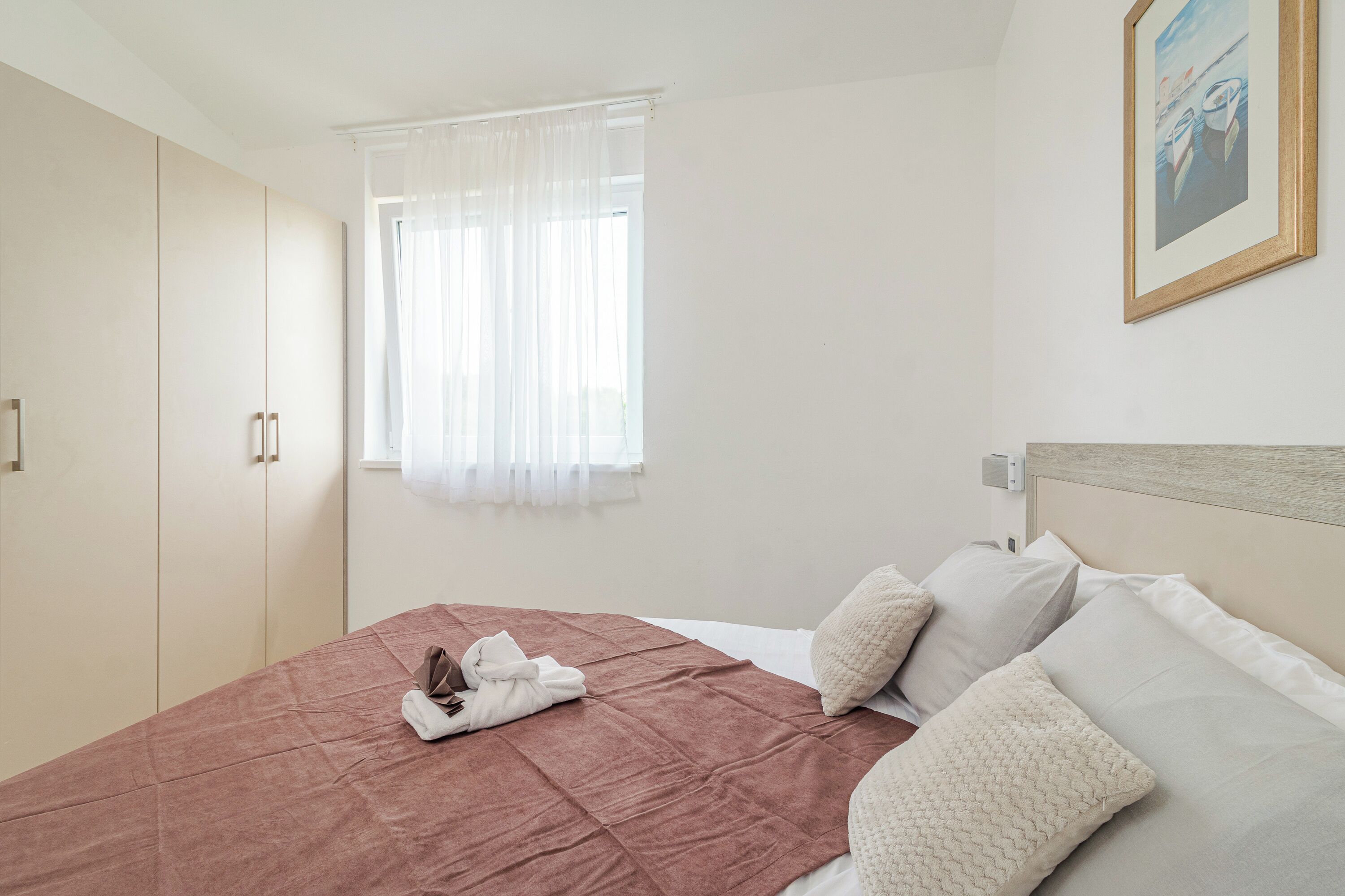 A bedroom with a bed, a window with sheer curtains, a painting on the wall, and a wardrobe.