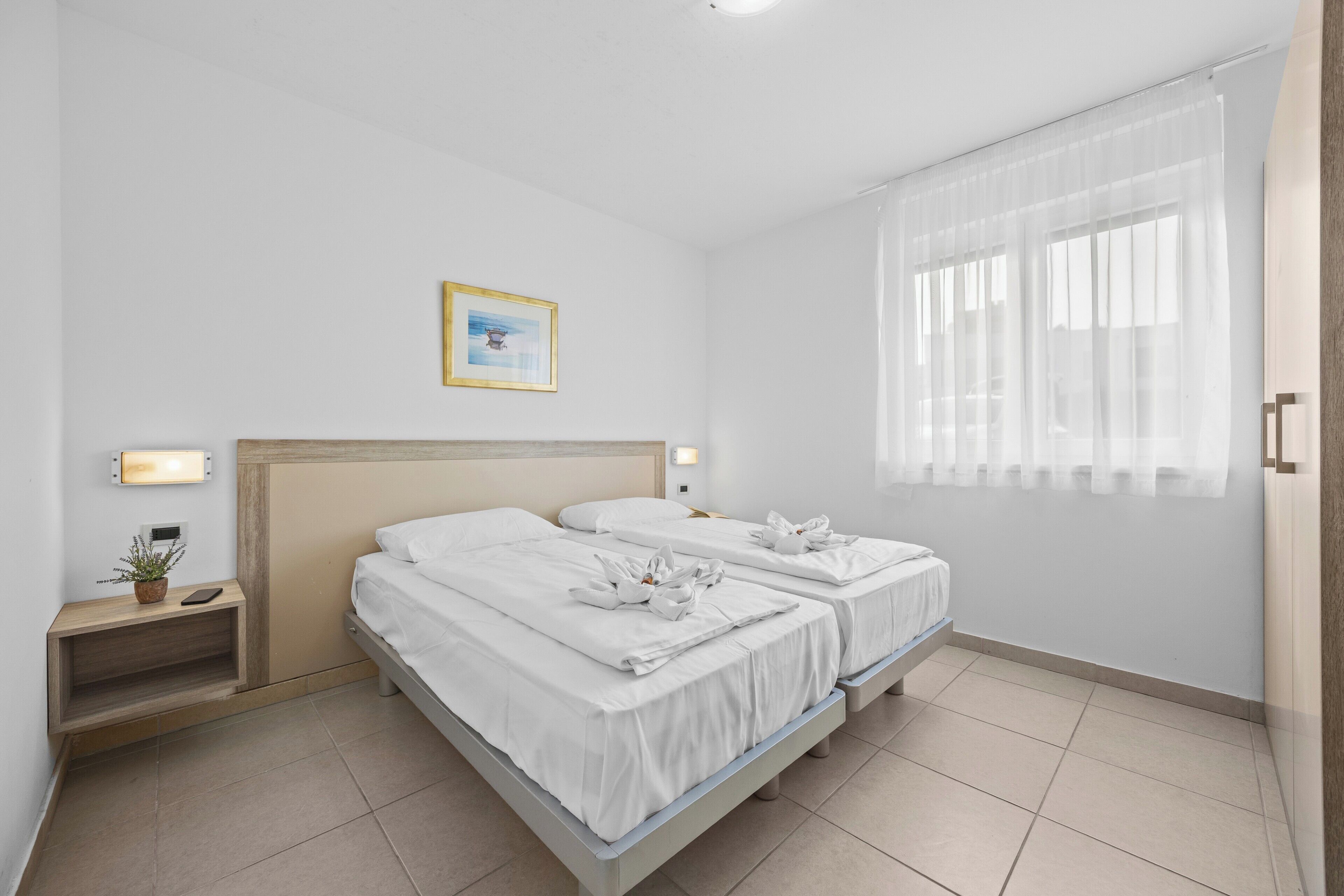 Ein Hotelzimmer mit zwei Betten, einem Nachttisch mit Lampe, einer kleinen Pflanze und einem Fenster mit transparenten Vorhängen.