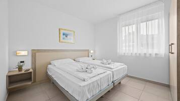 Habitación de hotel con dos camas, una mesita de noche con lámpara, una planta pequeña y una ventana con cortinas translúcidas.