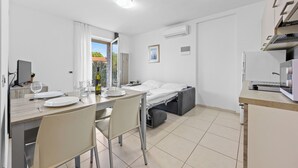 Una habitación compacta con una mesa de comedor preparada para dos personas, una cama con colchón gris, una pequeña cocina y una ventana con cortinas.