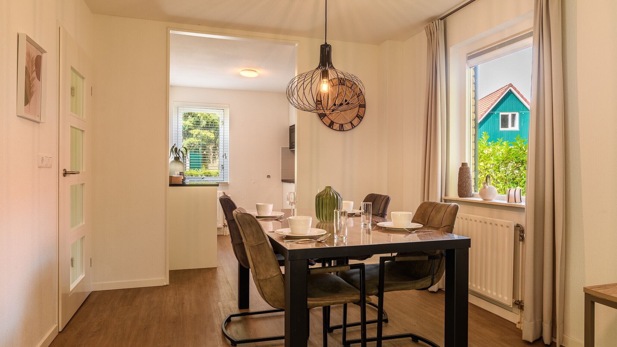 Un coin repas avec une table dressée pour quatre personnes, des chaises, un luminaire suspendu et une vue sur une maison bleue, visible à travers une fenêtre.