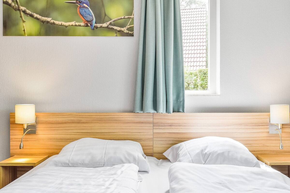 A hotel room with a wooden headboard, two bedside lamps, a window with curtains, and a wall-mounted picture of a bird.