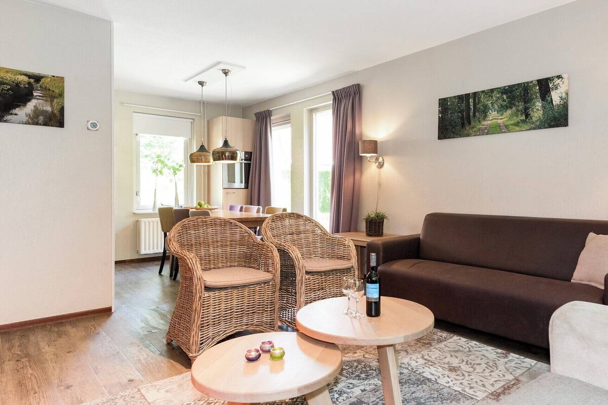 A living room with a brown sofa, wicker chairs, a small round table with a bottle and glasses, and a dining area with a table and chairs.