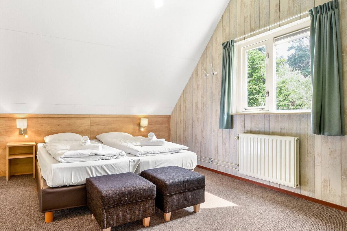 A bedroom with a sloped ceiling, a wooden headboard, a bed with white linens, two bedside tables with lamps, and a window with curtains.