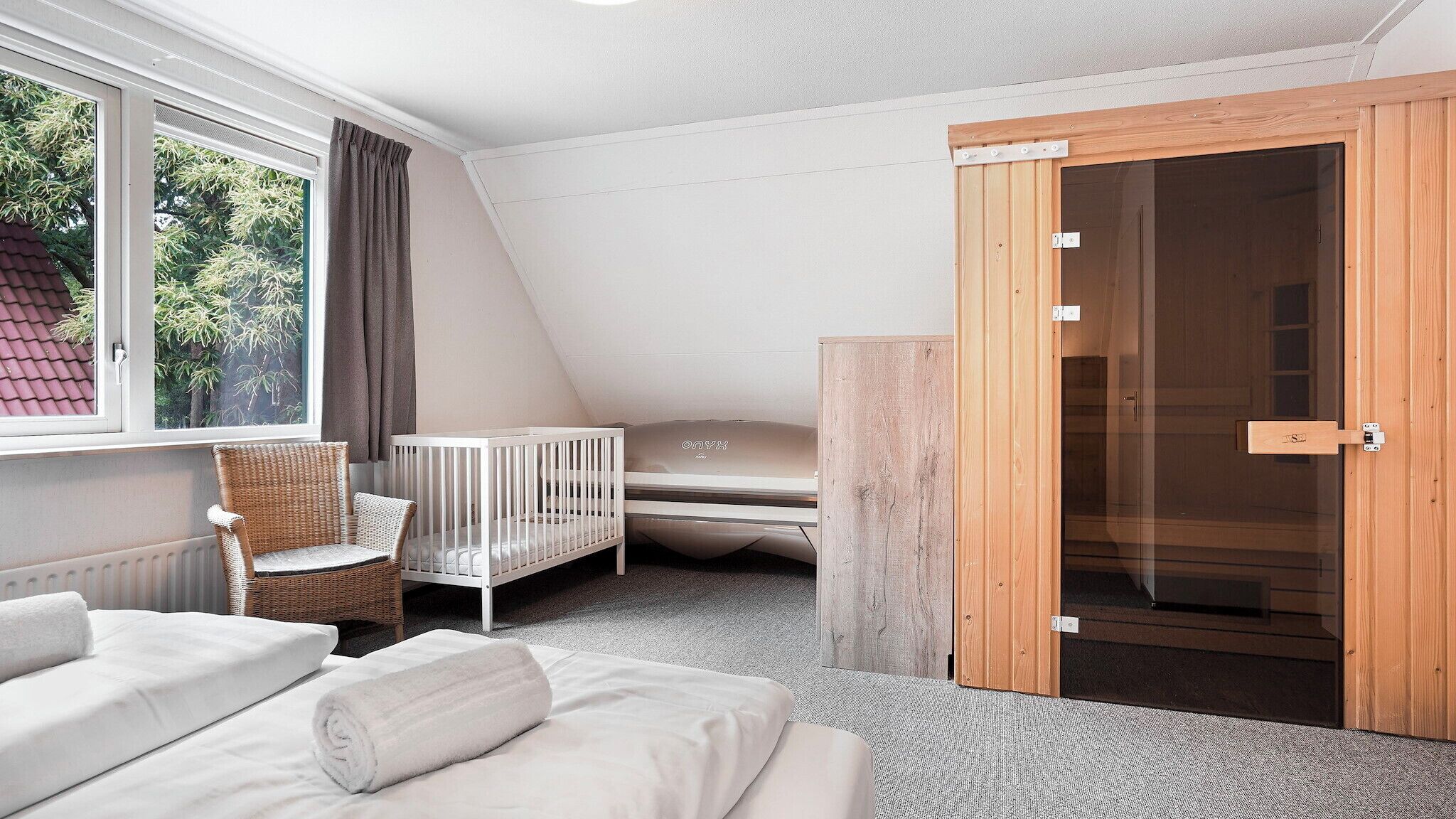 Un dormitorio con techo inclinado, una cuna, una silla de mimbre, una cama con ropa blanca y una puerta de madera.