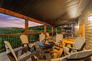A covered outdoor deck with Adirondack chairs, a stone fire pit, and a view of a forested landscape. - Private Pool! Rustic Charm! Steps from the Lake! (Branson)