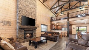 A spacious living area with a stone fireplace, leather sofas, a flat-screen TV, and a kitchen in the background. - Private Pool! Rustic Charm! Steps from the Lake! (Branson)