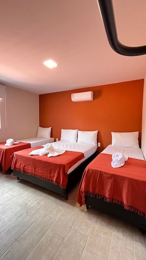 Habitación de hotel con dos camas, cada una con una colcha roja y almohadas blancas. Se observa un equipo de aire acondicionado instalado en la pared sobre las camas.