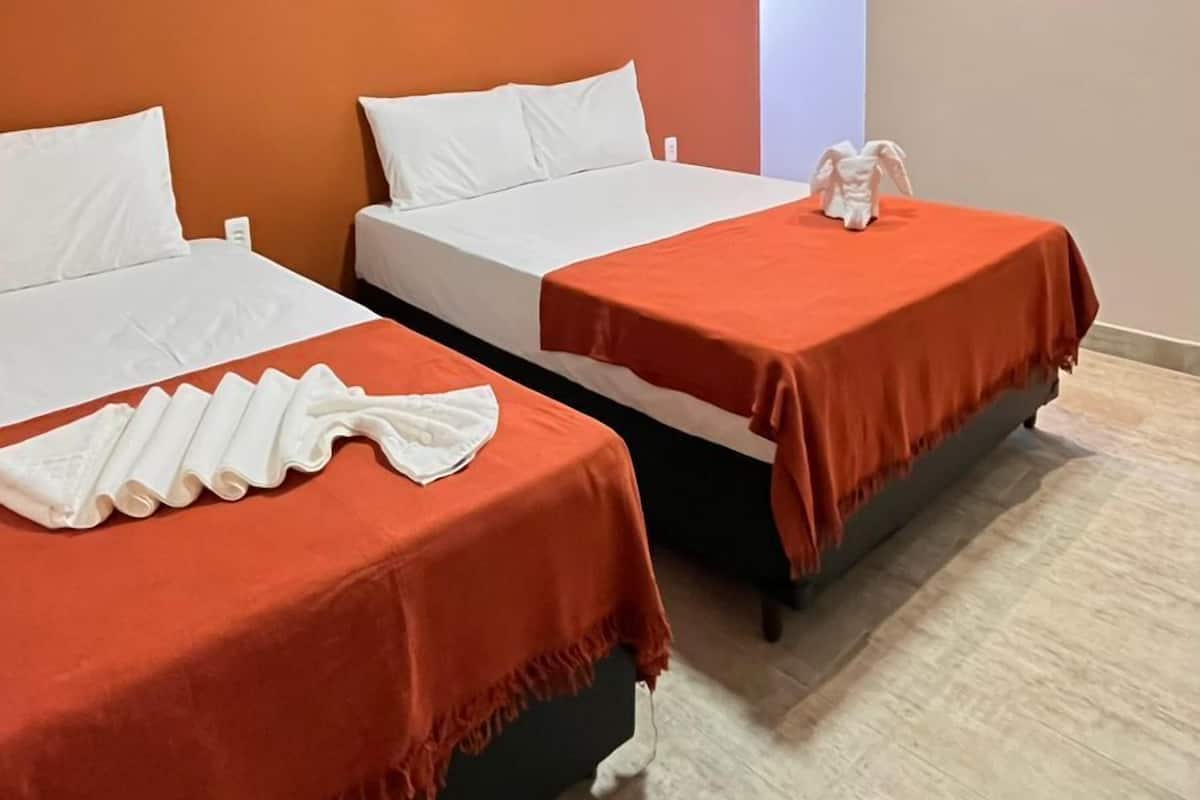Ein Hotelzimmer mit zwei Betten, jeweils mit weißer Bettwäsche und orangenen Bettdecken. Eine Klimaanlage ist an der Wand oberhalb der Betten angebracht.
