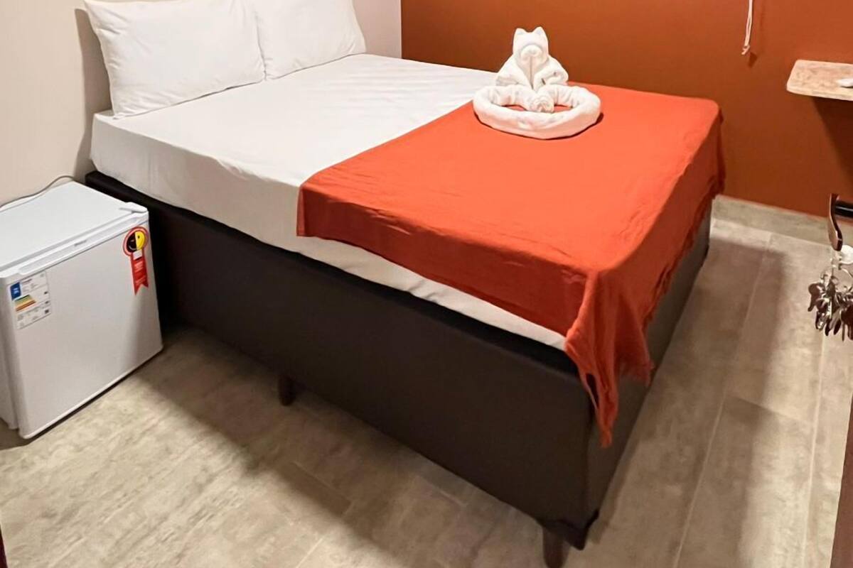 Ein Hotelzimmer mit einem Bett, einem kleinen Kühlschrank, einer Klimaanlage und einem Fenster mit einem weißen Rollo.