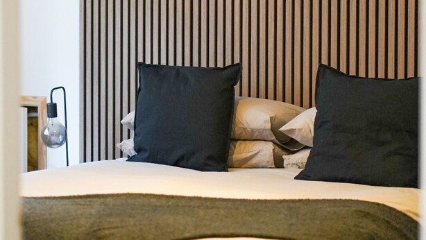 A bedroom with a bed, pillows, a bedside lamp, and a wall with vertical wooden slats. - No 3 @ The Rocket Rooms Apartments (Berwick-upon-Tweed)