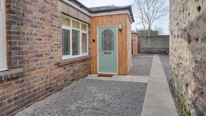 A brick building with a green door, a window with a decorative glass design, and a gravel pathway leading to a wooden structure. - No 3 @ The Rocket Rooms Apartments (Berwick-upon-Tweed)