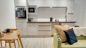 A modern kitchen with white cabinets, a microwave, and a toaster. There's a dining table with chairs and a green sofa with a remote control on it. - No 3 @ The Rocket Rooms Apartments (Berwick-upon-Tweed)