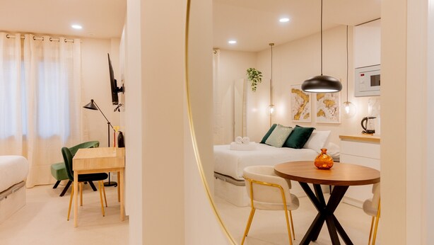 A modern hotel room with a dining area, a bed with a green pillow, a small table with a vase, and a wall-mounted TV.