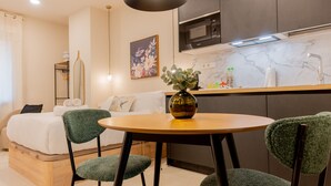 En kompakt opholdsstue med seng, et lille spisebord med stole, et tekøkken med mikroovn og en vase med blomster på bordet.