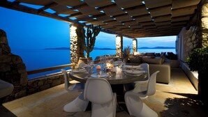 An outdoor dining area with a view of the sea, stone walls, and a pergola. - Topos Exclusive Mykonos  (Mykonos)