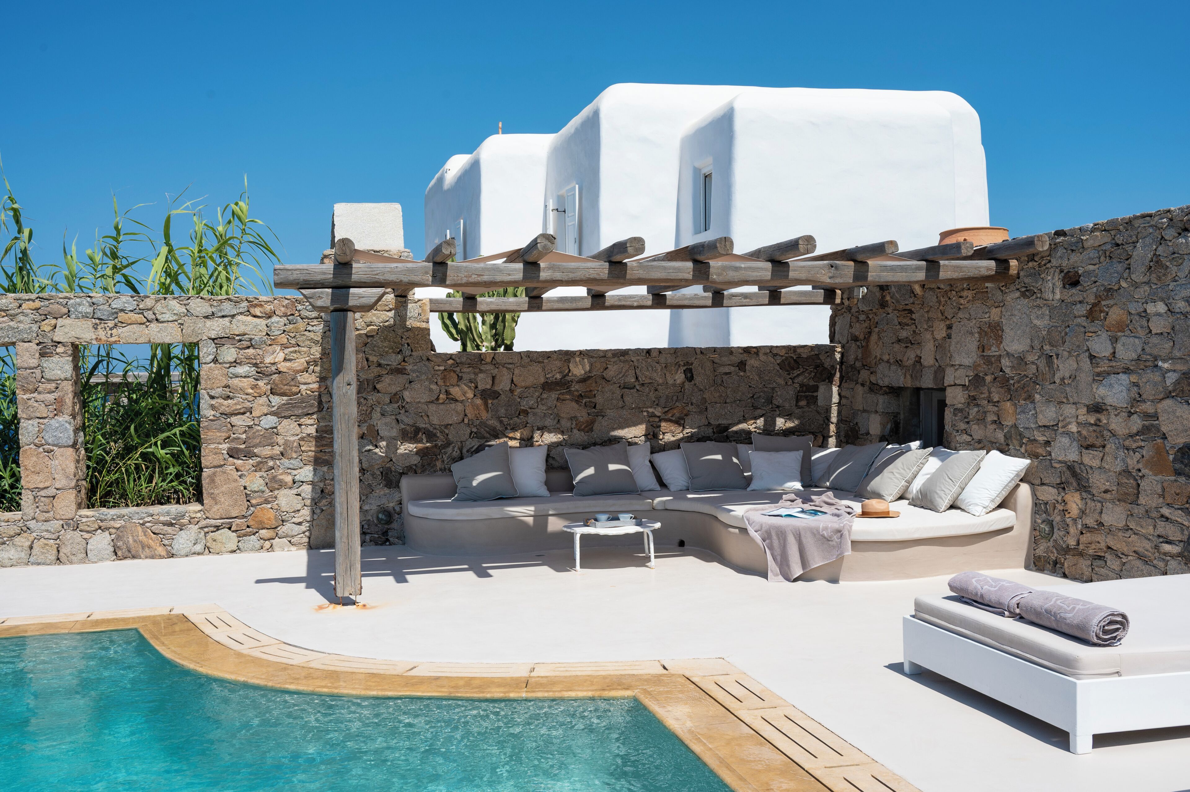 A white-washed building with a stone wall, a pool, and a wooden pergola.