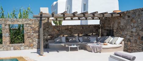 A white-washed building with a stone wall, a pool, and a wooden pergola.