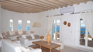 A spacious living room with a wooden ceiling, white sofas, a coffee table, and large windows with a view of the sea. - Topos Exclusive Mykonos  (Mykonos)