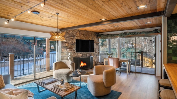 A cozy living room with a stone fireplace, comfortable seating, and large windows offering a view of the outdoors.