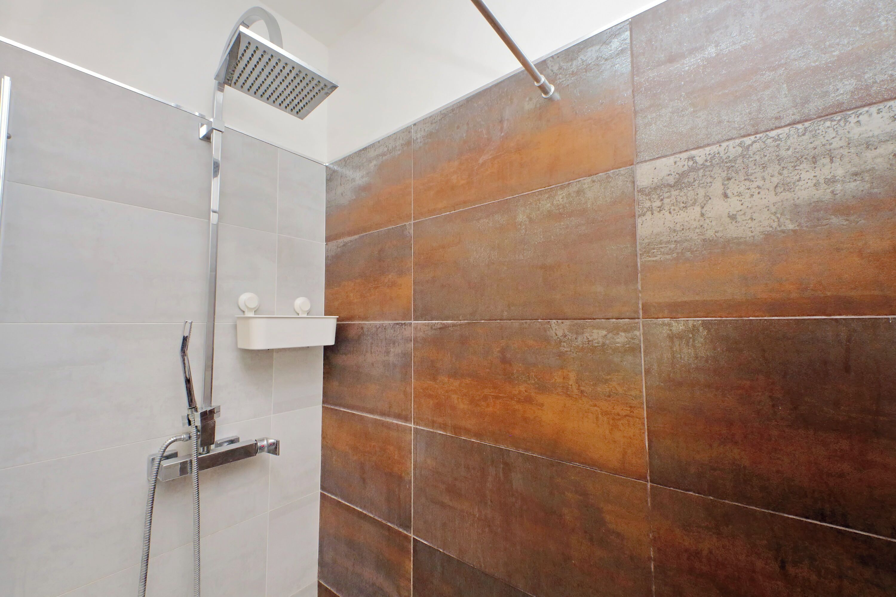 A shower area with a rain showerhead and a handheld showerhead, a white shelf, and a wall with large, rectangular tiles in shades of brown and beige.