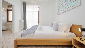 Ein Schlafzimmer mit einem hölzernen Bett, weißer Bettwäsche, einem Nachttisch, einer Lampe und einem durchgehenden Türspalt einsehbaren Badezimmer.