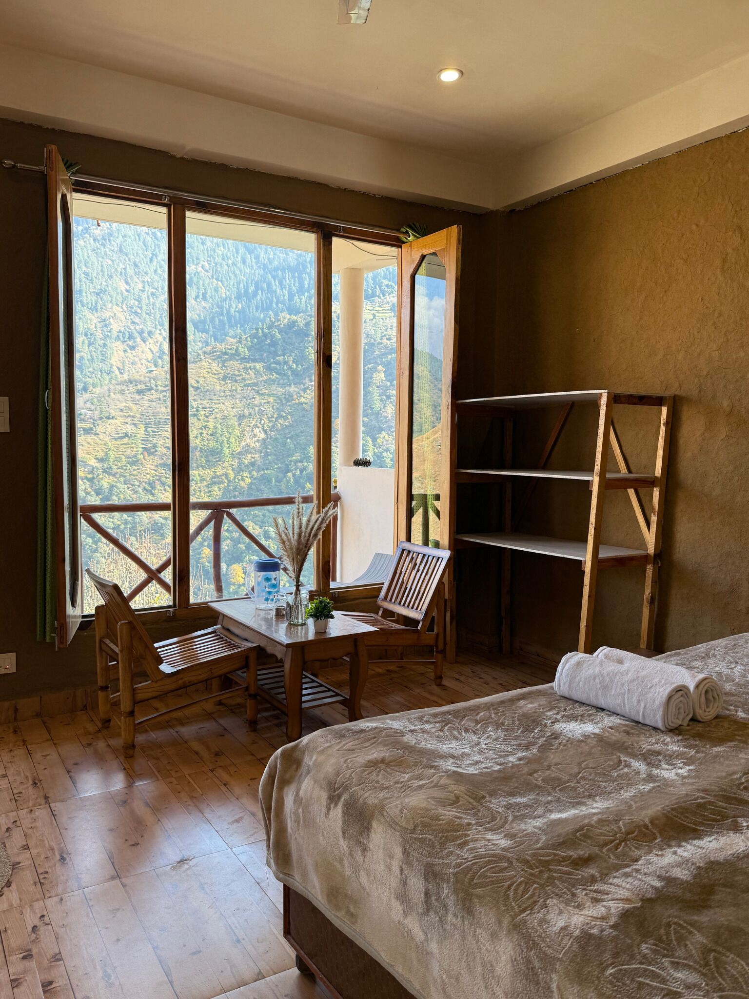 A room with a bed, a wooden table with chairs, a view of a mountainous landscape through large windows, and a wooden shelf unit.