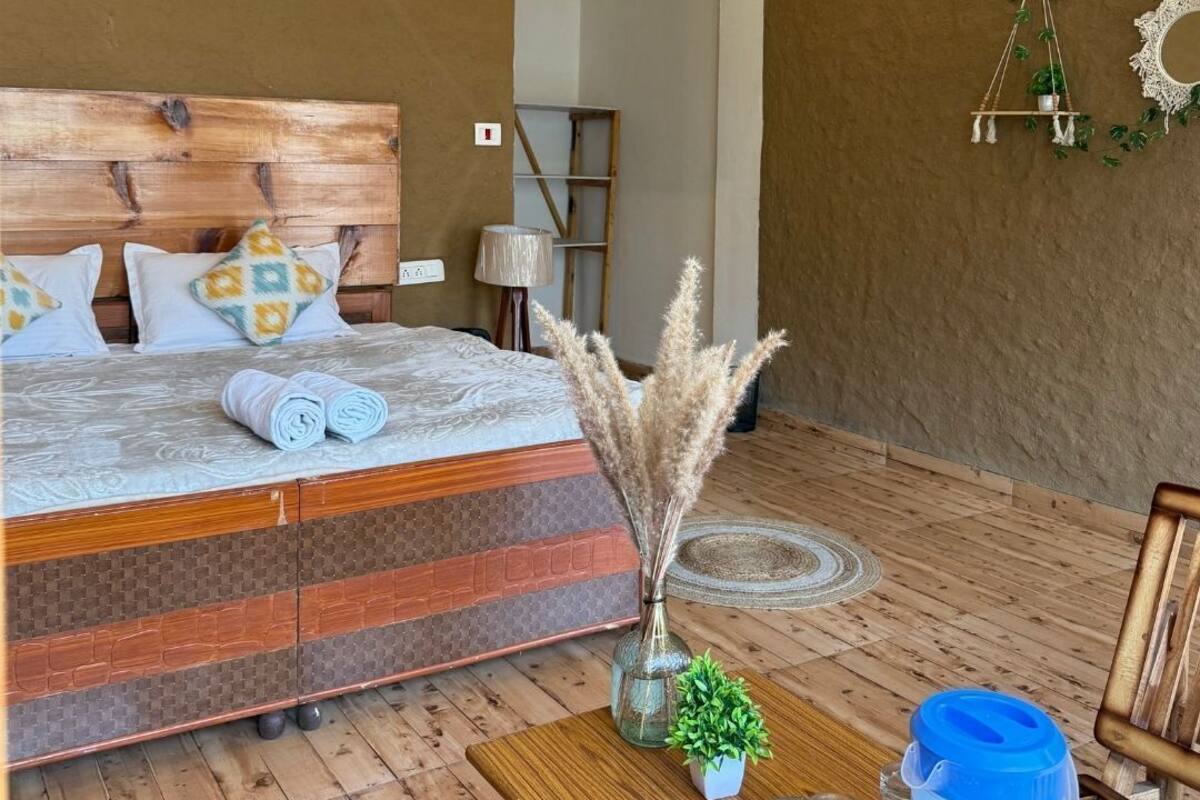Quarto com cabeceira de madeira, cama com roupa de cama estampada, mesa de cabeceira com uma planta, uma garrafa de água azul e um pequeno vaso com planta.