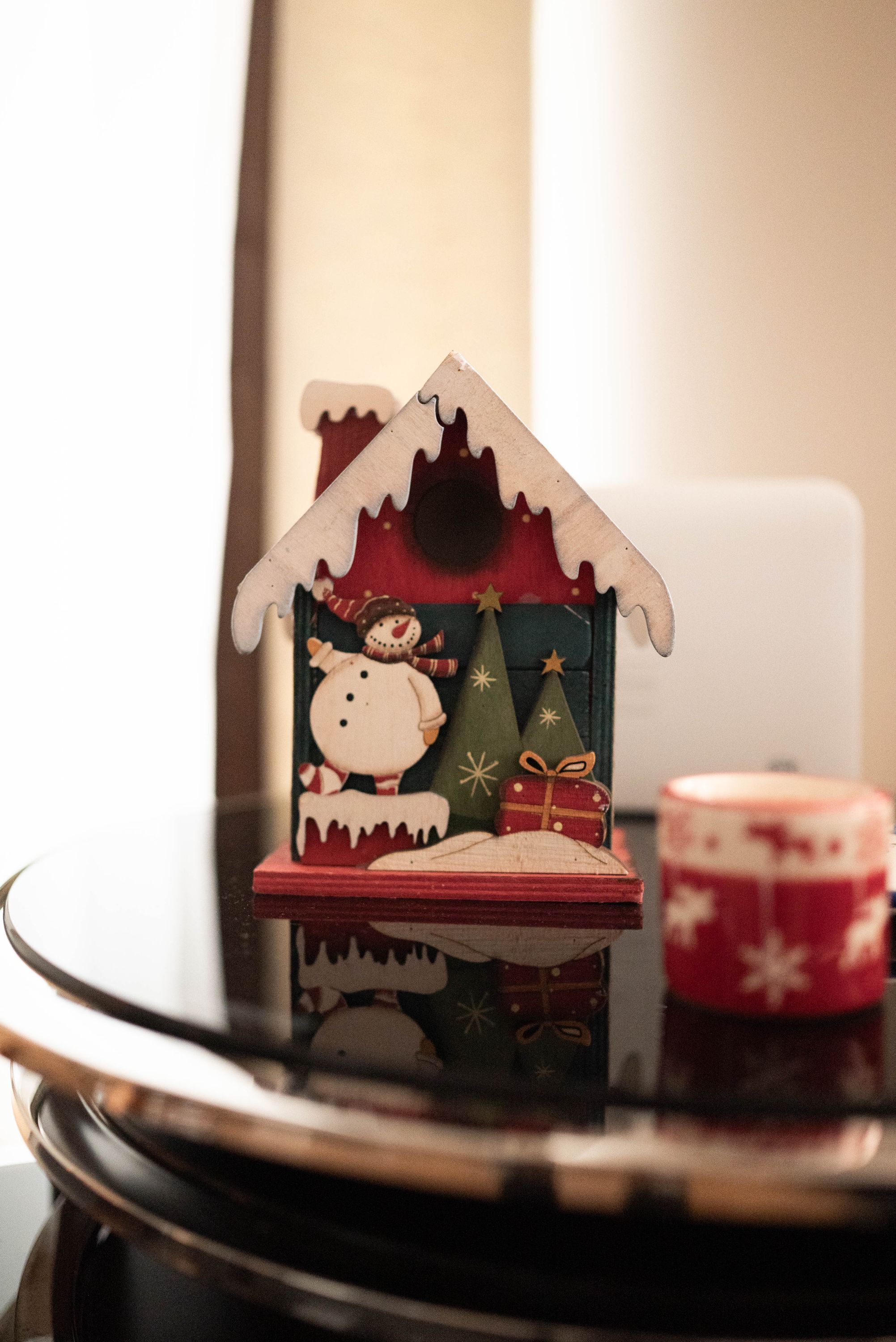 Une petite maison en bois, décorée d’un bonhomme de neige et de sapins de Noël, le tout reflété sur une surface brillante.