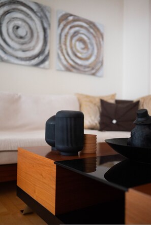 A modern living room with a wooden coffee table, a black speaker, and a stack of books. There is a white sofa with patterned cushions in the background. - Home Sweet Home Trikala Town (Trikala)