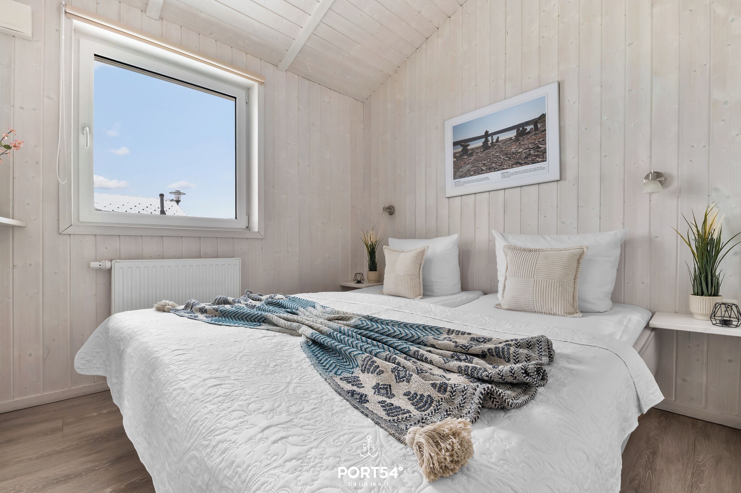 Ein Schlafzimmer mit großem Fenster, einem Bett mit weißer Bettwäsche, einer gemusterten Tagesdecke und einem gerahmten Bild an der Wand.