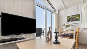 A modern room with a large flat-screen TV, wooden walls, a dining area with a table and chairs, and a window with a view of greenery. - Panorama Dream 47 - Scandinavian, cozy vacation home for 2 to 4 people (St. Andreasberg)