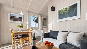Ein gemütlicher Raum mit Holzbalkendecke, einem Esstisch mit Stühlen, einer Couch, einer Orangenschale, einer Weinflasche und zwei gerahmten Bildern an den Wänden.