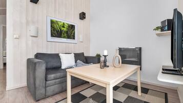 A modern living room with a grey sofa, a wooden coffee table, a framed picture on the wall, a television, and a small shelf with a plant.