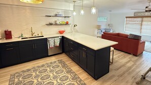 Une cuisine moderne avec des meubles de couleur sombre, un plan de travail clair et un lave-vaisselle encastré. Le salon est doté d’un canapé rouge et d’une fenêtre avec des stores.