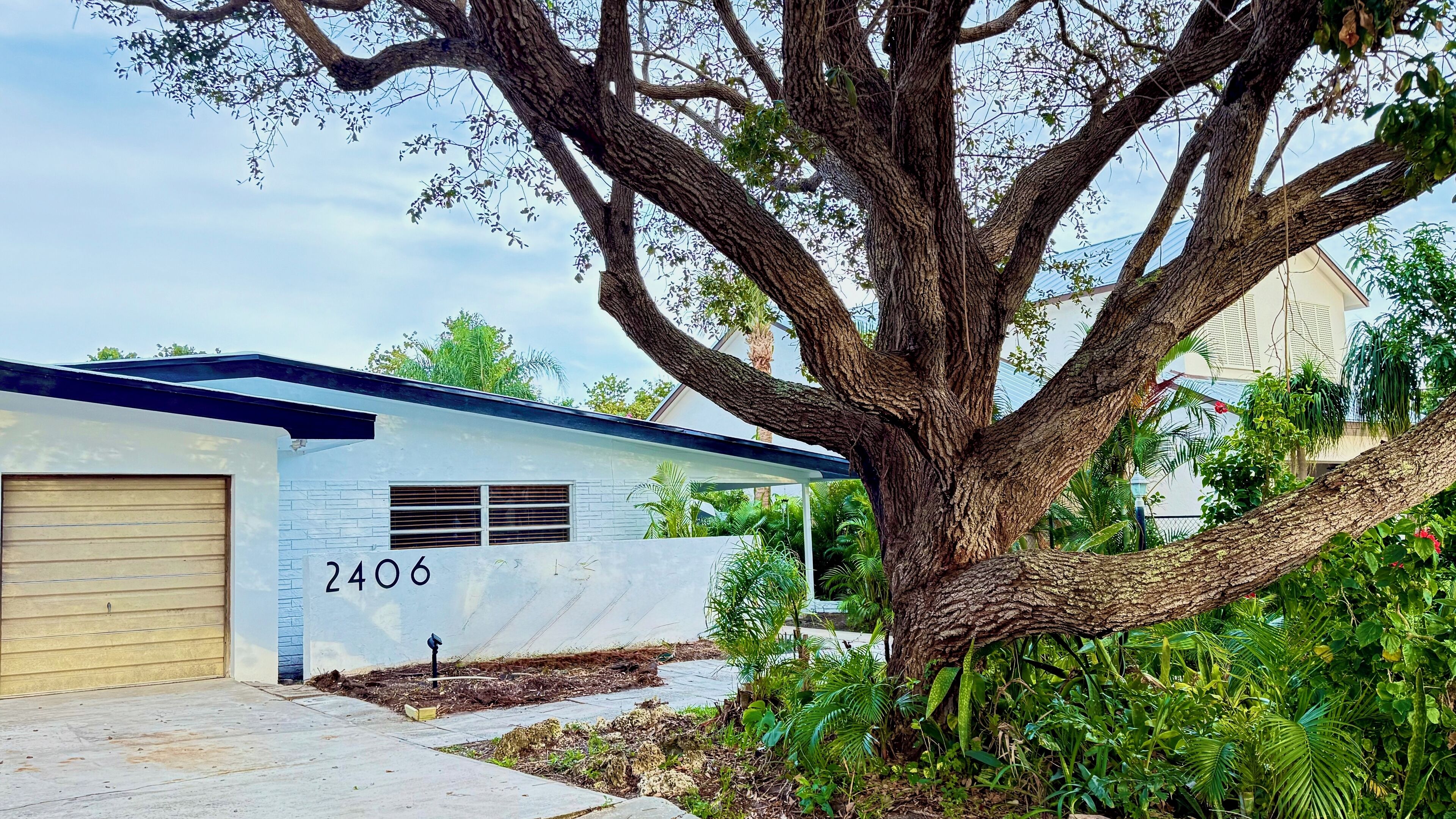 A modern house with a garage, number 2406, surrounded by lush greenery and a large tree.