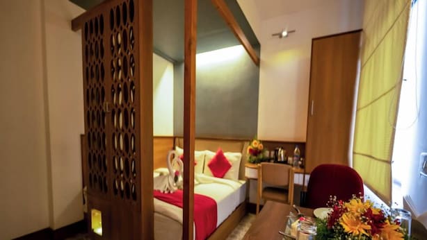 A hotel room with a bed, a desk with a chair, a wooden partition, and a bathroom visible through an open door.