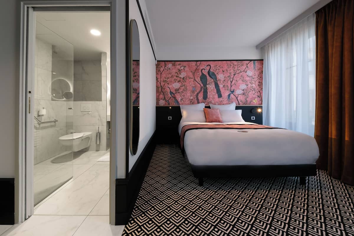 A modern hotel room with a large bed, a glass-enclosed shower area, and a decorative wall with floral and bird patterns.