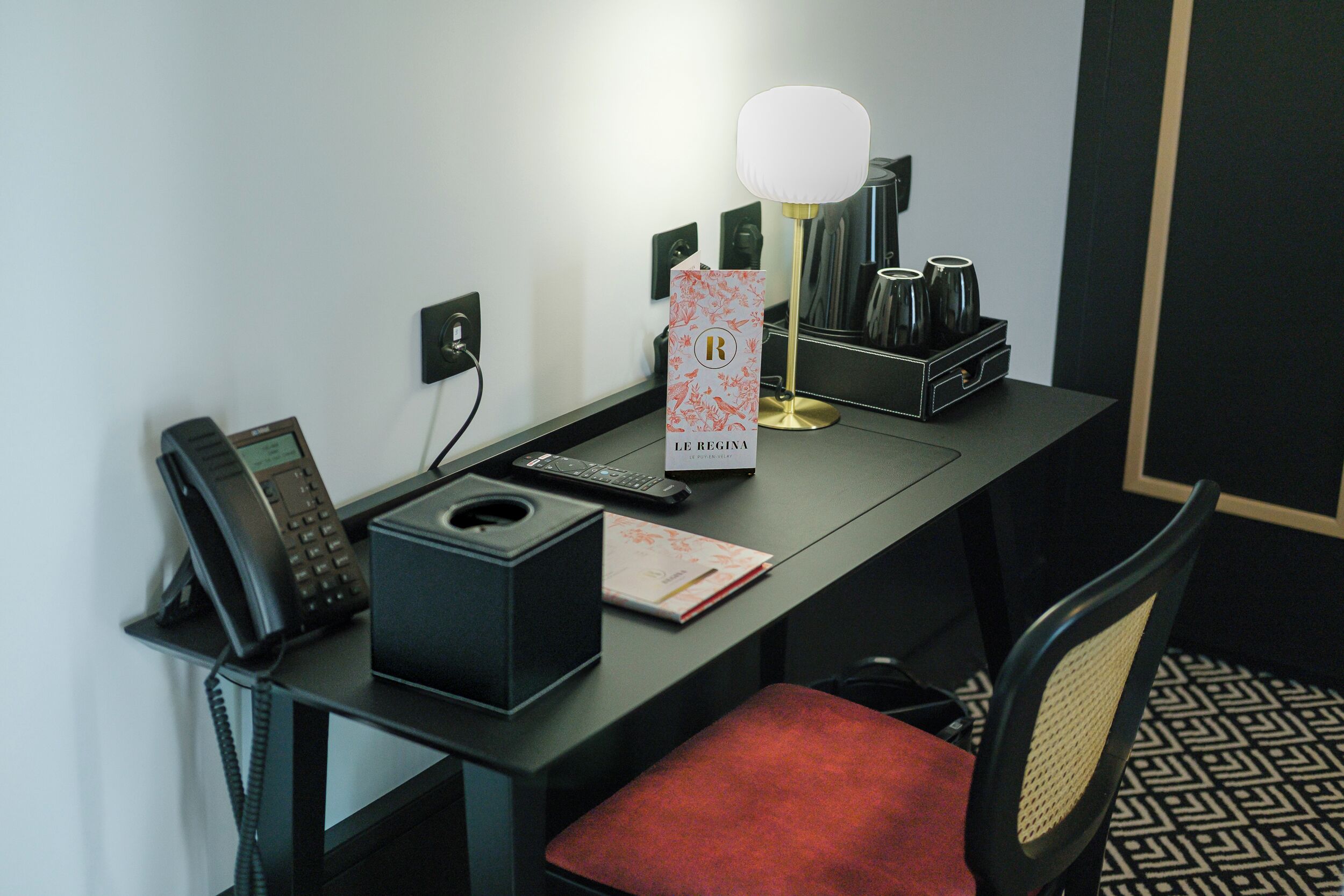 A hotel desk with a telephone, a lamp, a coffee maker, and a red chair.