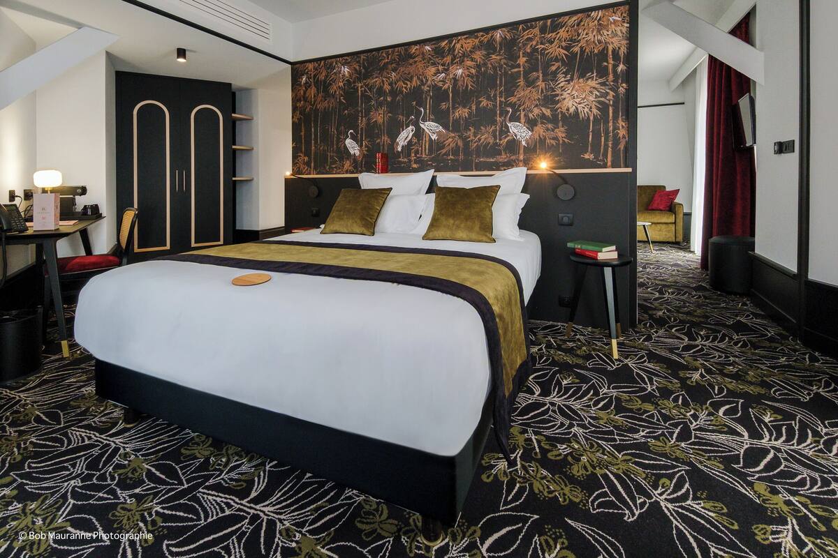 A modern hotel room with a large bed, a desk with a lamp, a bookshelf, and a wall with a nature-themed mural.