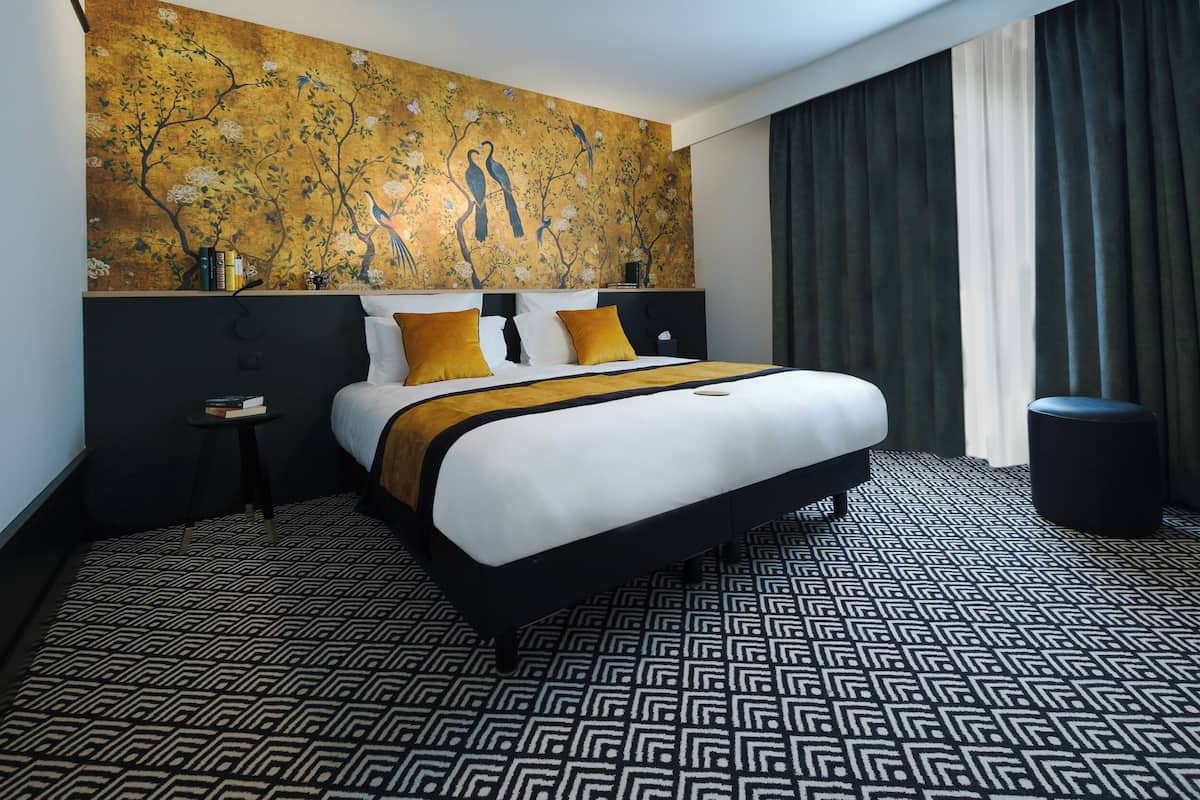 A hotel room with a large bed, a headboard with a floral mural, a small side table, and a patterned floor.