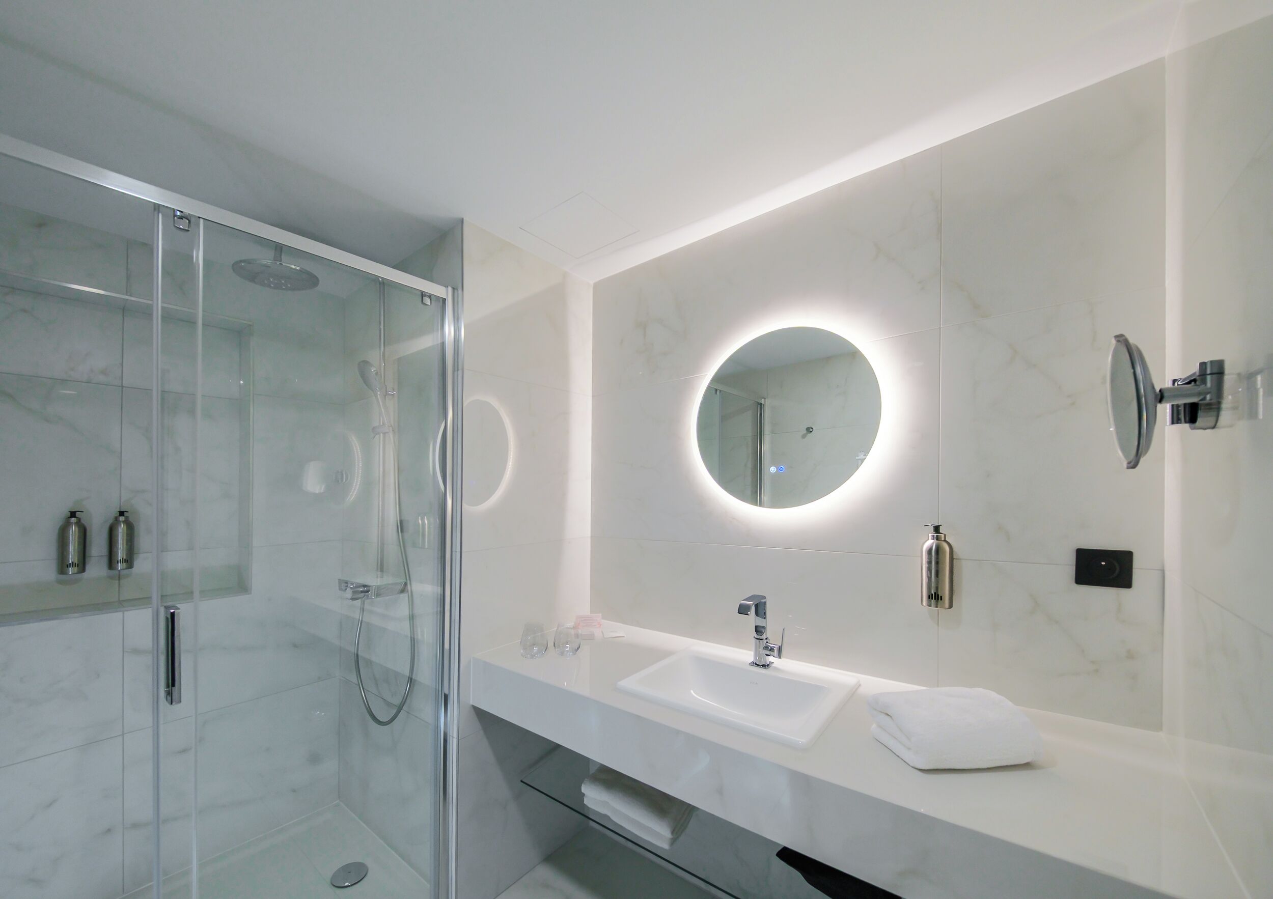 Une salle de bain moderne avec une douche à l’italienne en verre, un grand miroir avec un éclairage circulaire et un lavabo blanc.