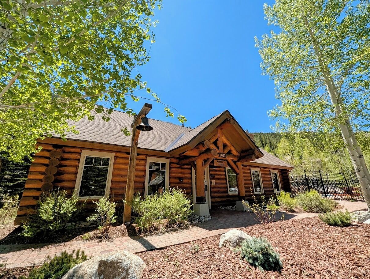 A log cabin with a porch, surrounded by trees and a clear sky.
