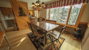 Zona pranzo con tavolo in legno, sedie e lampadario. Sullo sfondo, una cucina con pensili in legno e una finestra che mostra un bosco innevato.