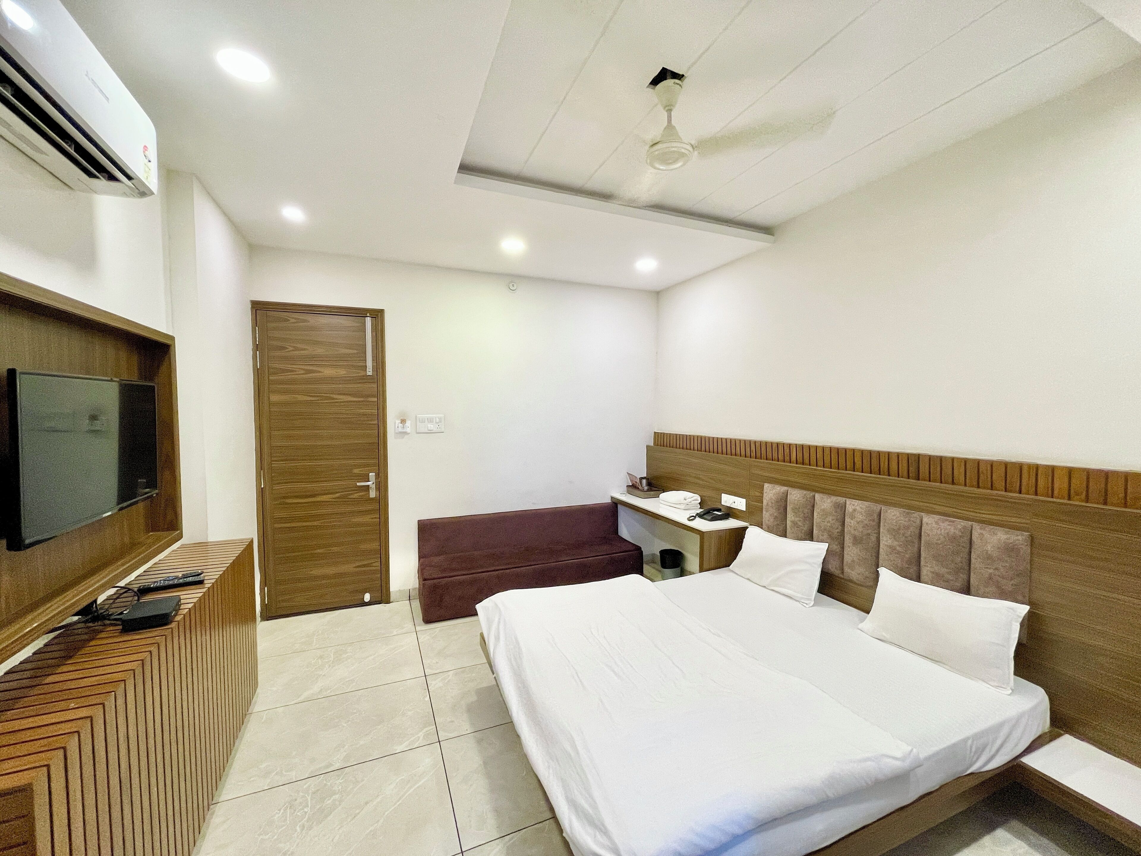 Quarto de hotel com cama, televisão presa a um painel de madeira, escrivaninha com cadeira e ventoinha no teto.